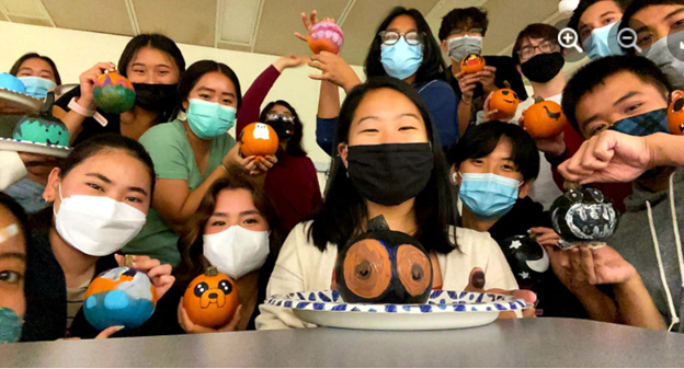 A group of students (around 15) all wearing masks hold up their painted pumpkins