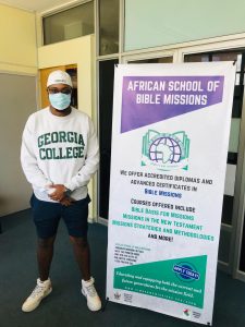 Student wearing mask and Georgia College sweatshirt standing next to sign reading African School of Bible Missions