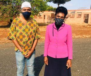 Man wearing a backwards baseball hat and wearing a mask standing next to woman in a pink blazer also wearing a mask in front of a school